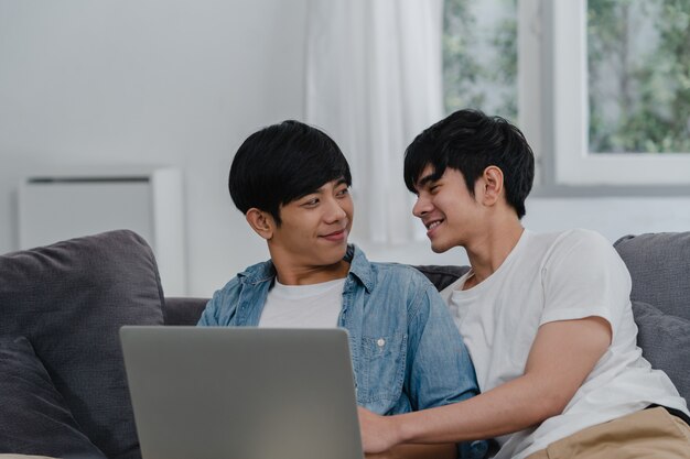 Young Gay couple using computer laptop at modern home. Asian LGBTQ+ men happy relax fun using technology watching movie in internet together while lying sofa in living room at house .