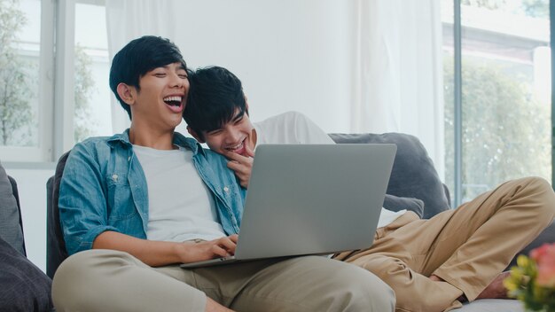 현대 집에서 컴퓨터 노트북을 사용하는 젊은 게이 커플. 아시아 LGBTQ + 남성은 집에서 거실에 소파에 누워있는 동안 인터넷에서 함께 영화를보고 기술을 사용하여 즐거운 휴식을 취하십시오.