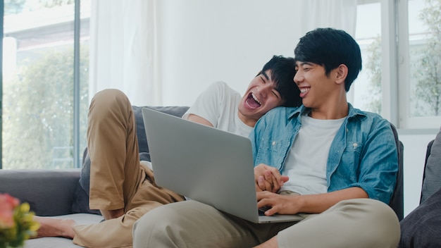 Young Gay couple using computer laptop at modern home. Asian LGBTQ+ men happy relax fun using technology watching movie in internet together while lying sofa in living room at house .