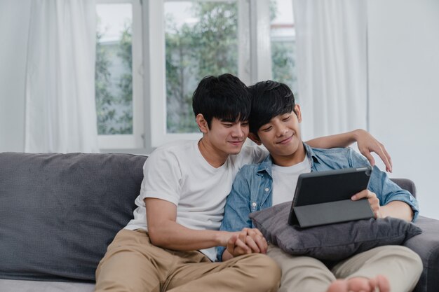 Young Gay couple using tablet at home. Asian LGBTQ+ men happy relax fun using technology watching movie in internet together while lying sofa in living room .