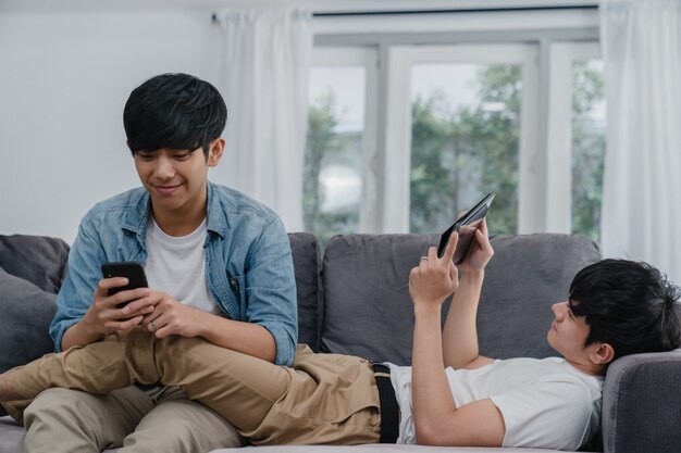 現代の家で携帯電話とタブレットを使用して若いゲイlgbtqカップル。アジアの恋人男性幸せなリラックスした笑いと楽しい技術は、リビングルームでソファに横たわっている間一緒にインターネットでゲームをプレイします。