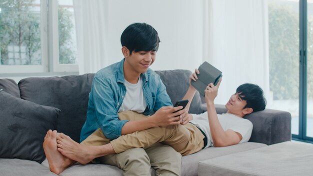 Young gay lgbtq couple using mobile phone and tablet at modern home. Asian lover male happy relax laugh and fun technology play games in internet together while lying sofa in living room .