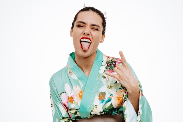 Молодая гейша в традиционном японском кимоно счастлива и взволнована, высунув язык, показывая рок-символ на белом
