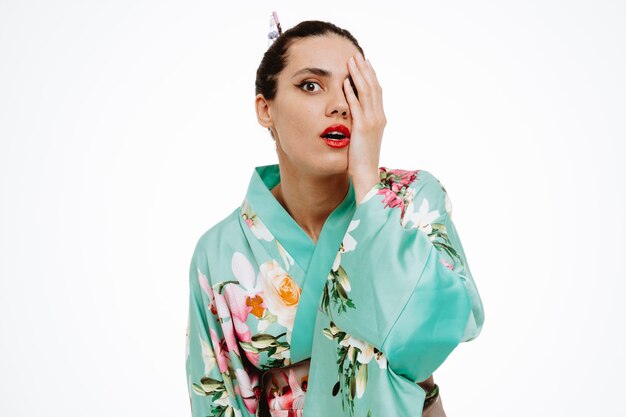 Young geisha woman in traditional japanese kimono amazed and surprised covering one eye with hand on white