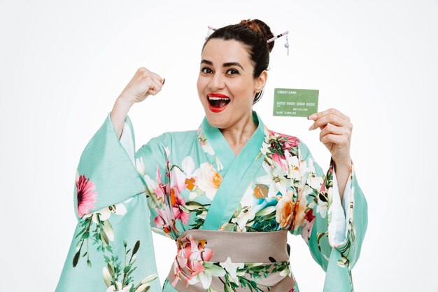 young geisha woman in traditional japanese kimono holding credit card clenching fist crazy happy and excited standing over white wall