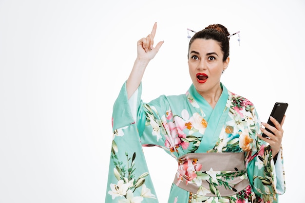 young geisha woman in traditional japanese kimono holding smartphone looking at front being surprised with smile on smart face pointing with index finger up have a good idea