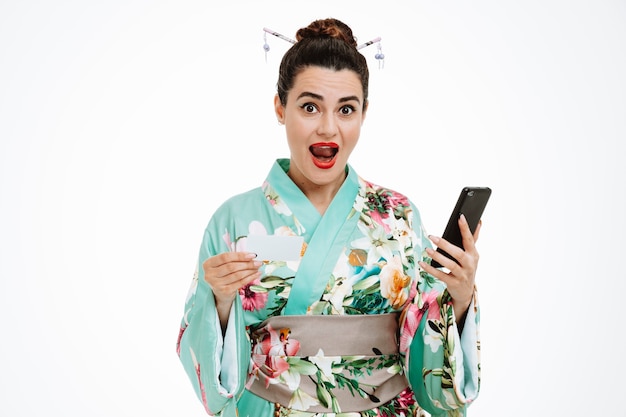 スマートフォンを保持している伝統的な日本の着物の若い芸者の女性は、白い壁の上に広く立って笑顔で幸せで幸せなクレジットカードを示しています