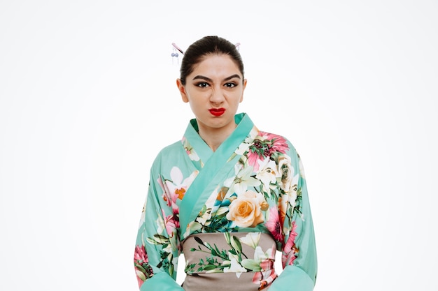 young geisha woman in traditional japanese kimono looking at front with angry face making wry mouth standing over white wall
