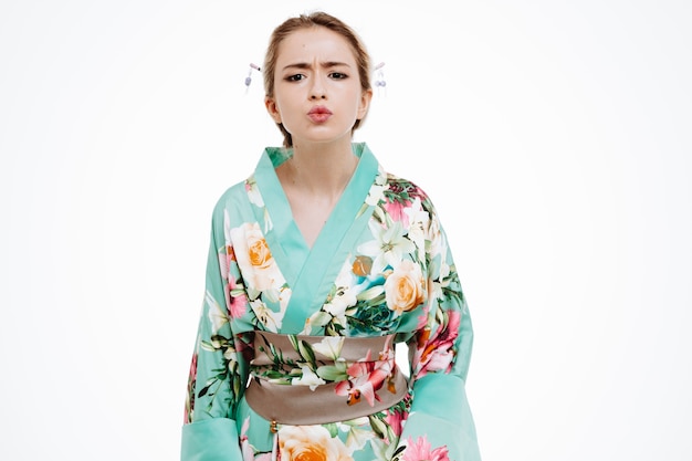 young geisha woman in traditional japanese kimono looking at front with frowning face standing over white wall