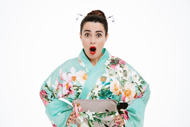 young geisha woman in traditional japanese kimono looking at front with wide open mouth and eyes being amazed and surprised standing over white wall