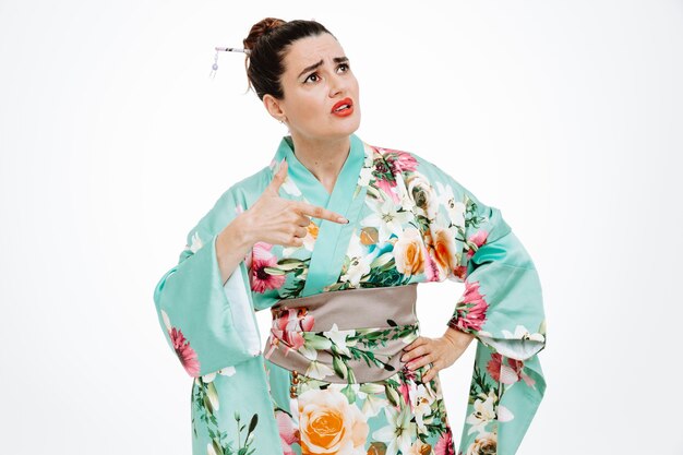 young geisha woman in traditional japanese kimono looking up puzzled pointing with index finger to the side standing over white wall
