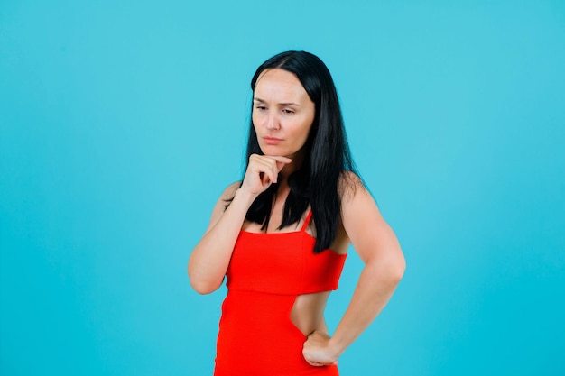 Young gir is thinking by holding hand on chin and putting other hand on waist on blue background