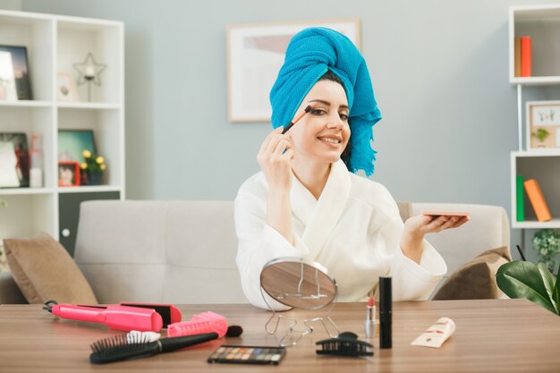 young girl applying eyeshadow with makeup brush wrapped hair in towel sitting at table with makeup tools in living room