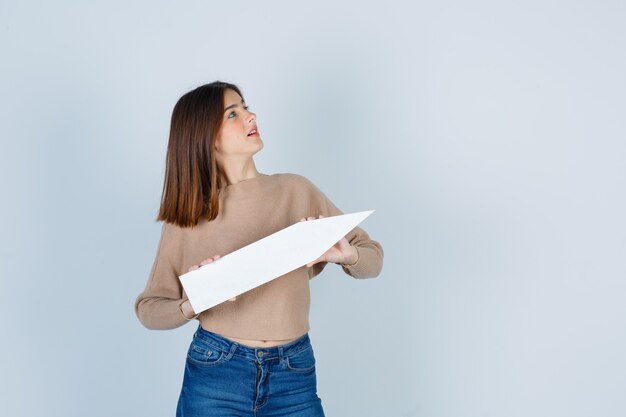 Young girl in beige knitwear, jeans holding paper stick, looking at right side and looking cute , front view.