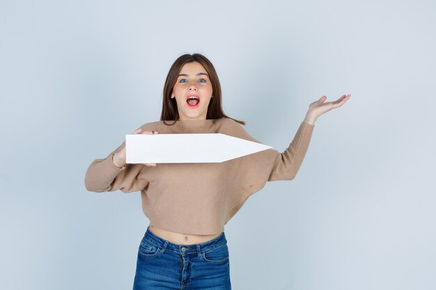 Young girl in beige knitwear, jeans holding paper stick, spreading palm out and looking surprised , front view.