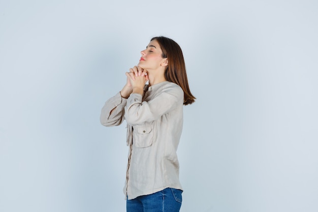 Young girl in beige shirt, jeans clasping hands in praying position and looking hopeful , front view.