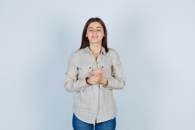 Young girl in beige shirt, jeans holding one hand on arm, keeping eyes closed and looking cheerful , front view.