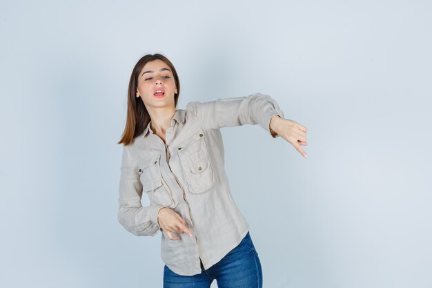 Young girl in beige shirt, jeans pointing down with index fingers and looking focused , front view.
