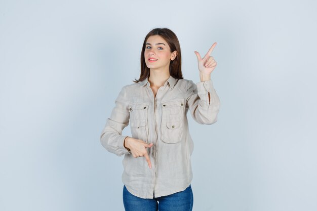 Young girl in beige shirt, jeans pointing up and down with index fingers and looking cute , front view.