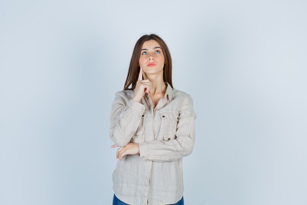 Young girl in beige shirt, jeans putting index finger on jowl, thinking about something and looking pensive , front view.