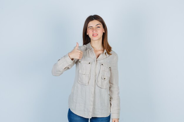 Молодая девушка в бежевой рубашке, джинсах показывает палец вверх, подмигивает и выглядит мило, вид спереди.