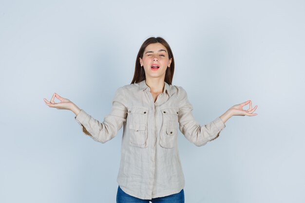 Young girl in beige shirt, jeans standing in meditating pose, winking and looking cheery , front view.