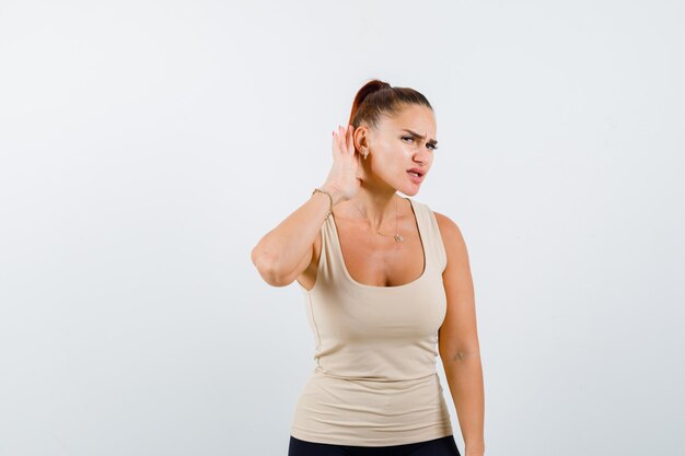 Young girl in beige top, black pants holding hands near ear to hear something and looking focused , front view.