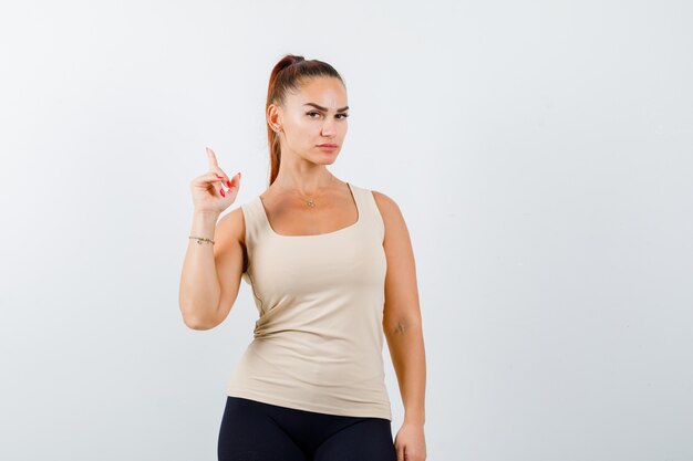 Young girl in beige top, black pants raising index finger in eureka gesture and looking sensible , front view.