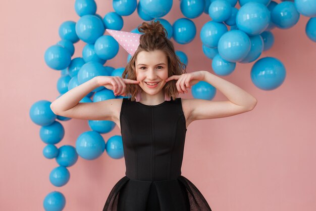 young girl in a birthday hat posing holding fingers on cheeks at pink wall and blue balloons