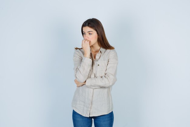 Young girl biting fist in beige shirt, jeans and looking pensive. front view.