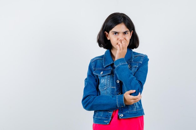 Young girl biting fist while holding one hand on elbow in red t-shirt and jean jacket and looking excited , front view.