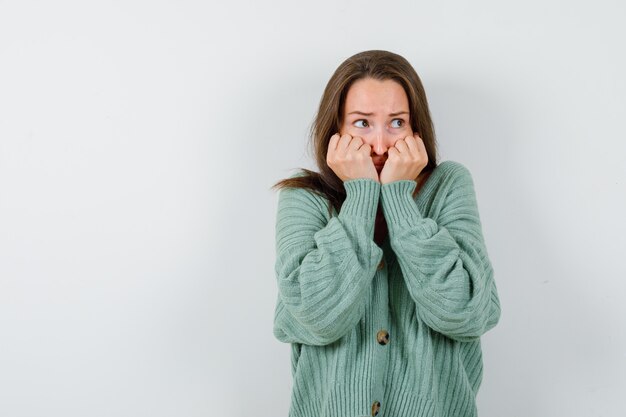 Young girl biting fists emotionally, looking away in knitwear, skirt and looking scared , front view.