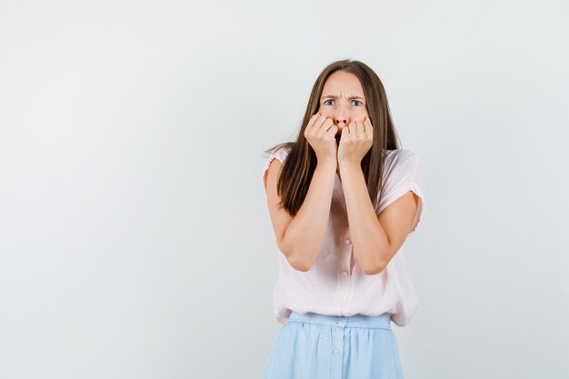 Young girl biting fists emotionally in t-shirt, skirt and looking scared , front view.