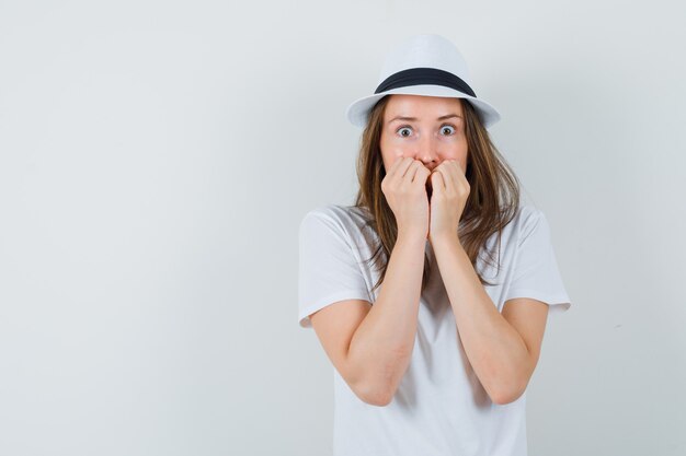 Young girl biting fists emotionally in white t-shirt, hat and looking frightened. front view.