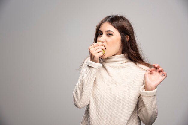 A young girl biting a green apple on a gray wall.