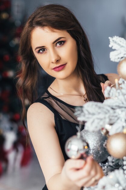 Young girl in a black dress poses before a shiny Christmas tree