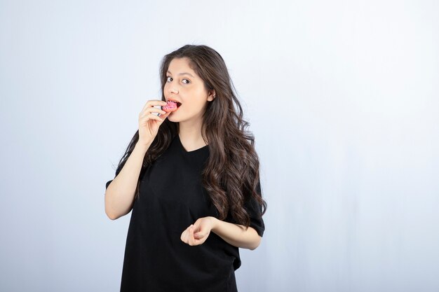 Young girl in black outfit eating pink cookie on white wall . 