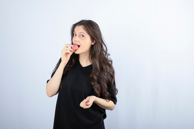 Молодая девушка в черном наряде ест розовое печенье на белой стене.