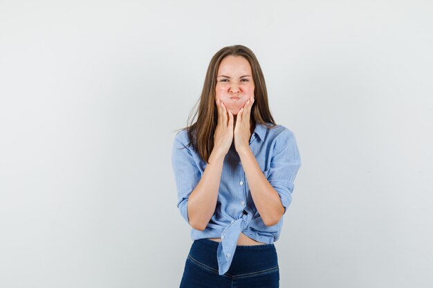 Young girl blowing cheeks with fingers on jaw in blue shirt, pants and looking gloomy