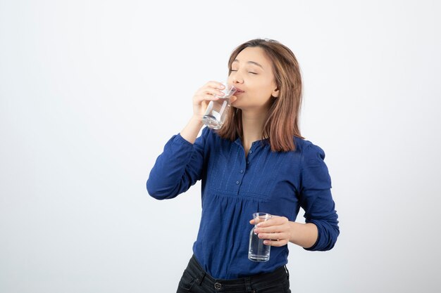 young girl in blue blouse drinking glass of water on white wall.