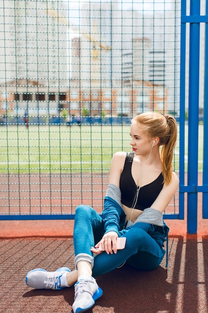 黒のトップと青いスポーツスーツの少女は、スタジアムのフェンスの近くに座っています。彼女はヘッドフォンで音楽を聴いています。彼女は魅力的な人物です。