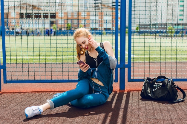 Молодая девушка в синем спортивном костюме с черным верхом сидит у забора на стадионе. Она слушает музыку в наушниках и улыбается телефону.