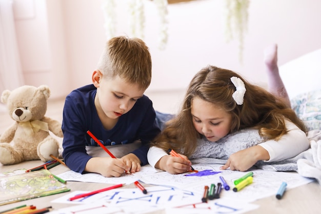A young girl and a boy are drawing in coloring books lying in the room on the floor