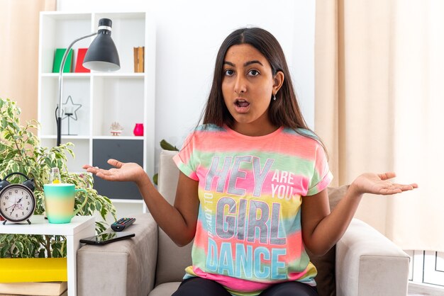 Young girl in casual clothes  confused spreading arms to the sides sitting on the chair in light living room