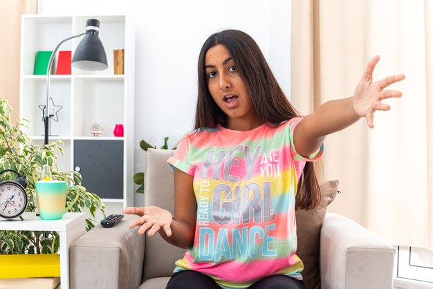 Young girl in casual clothes  confused and surprised spreading arms to the sides sitting on the chair in light living room