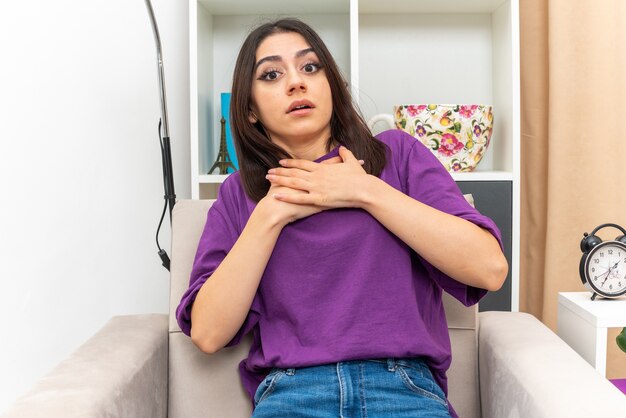 Young girl in casual clothes  confused and worried holding hands on her chest sitting on a chair in light living room