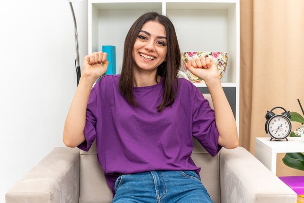 Young girl in casual clothes  happy and excited clenching fists sitting on a chair in light living room