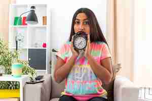 Free photo young girl in casual clothes holding alarm clock  surprised sitting on the chair in light living room