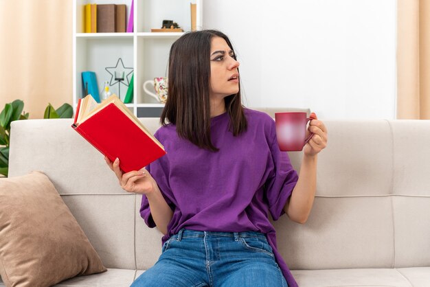 Young girl in casual clothes holding book and cup of tea looking aside puzzled sitting on a couch in light living room