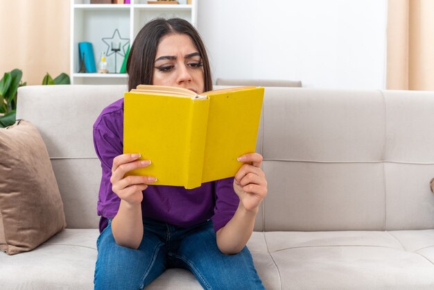 Молодая девушка в повседневной одежде держит книгу и читает с серьезным лицом, сидя на диване в светлой гостиной